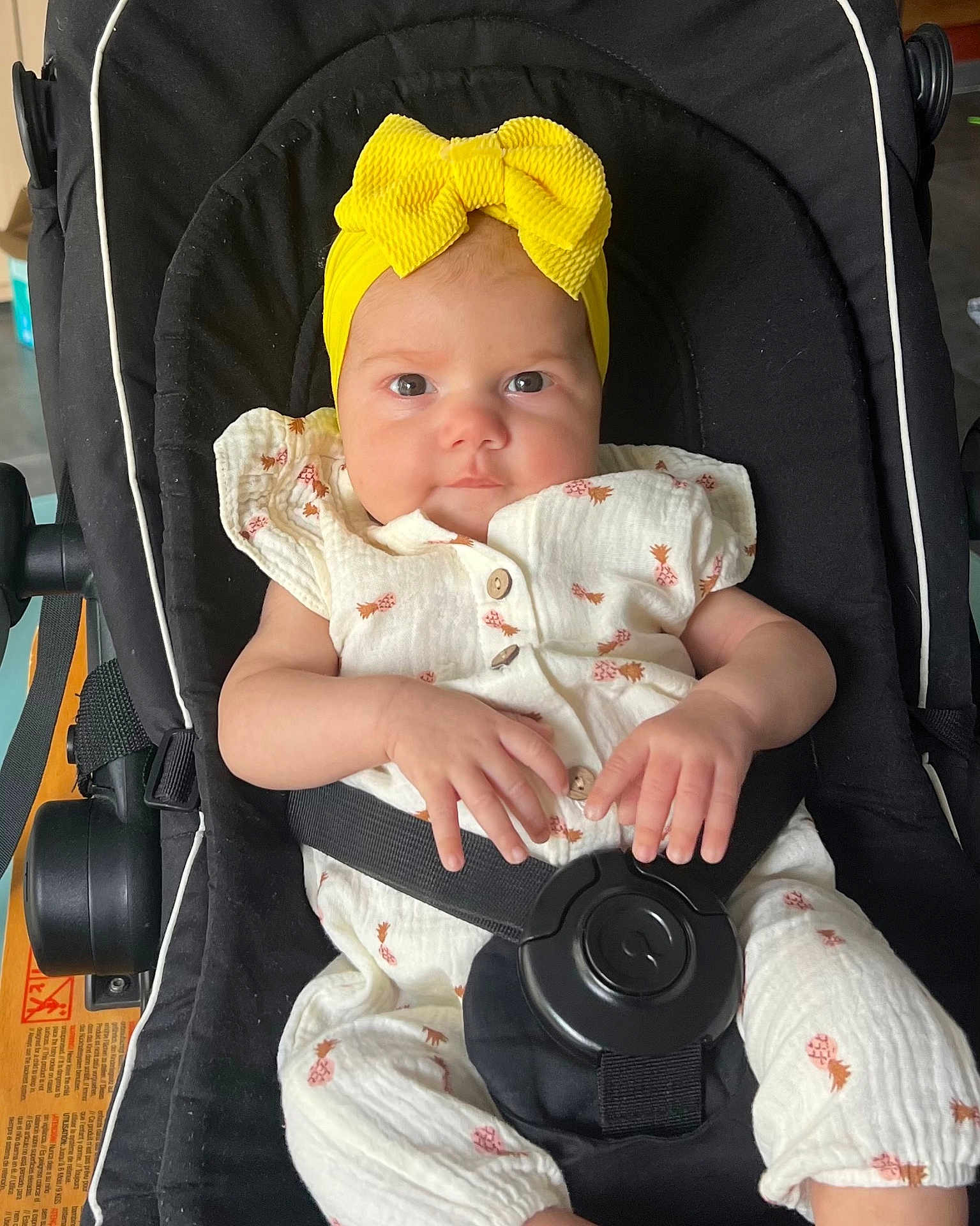 Olympe participe au concours pour gagner de l'argent avec cette photo : baby, child, infant, stroller, yellow_bow, headband, white_outfit, pineapple_pattern, black_stroller, seatbelt, hand, face, eyes, cute, indoors, person, portrait, sitting, clothing, soft_lighting