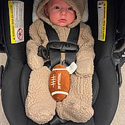 Deacon is registered to the contest to win money with this photo: baby, infant, child, car_seat, football, bear_suit, hood, 冬服, safety_harness, seated, portrait, indoor, wood_floor, black_car_seat, fluffy_clothing, cute, face, looking_at_camera, infant_carrier, photography