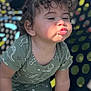 toddler, child, curly_hair, green_outfit, dinosaur_print, pouty_face, playground, kneeling, hand, fingers, outdoor, daylight, metal_structure, colorful_background, patterned_clothing, cute_expression, baby, person, fun, play