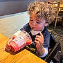 toddler, child, curly_hair, high_chair, smoothie, drink, straw, table, wooden_table, indoor, restaurant, casual, happy, cute, clothing, sitting, person, food, beverage