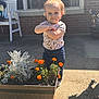 toddler, child, flower_planter, orange_flowers, plant, outdoor, sunlight, brick_wall, window, concrete, jeans, shirt, person, young_child, arms_crossed, shadow, garden, casual_clothing, daytime, smiling