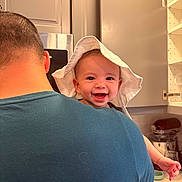Hudson is registered to the contest to win money with this photo: baby, child, person, smile, happy, hat, white_hat, kitchen, cabinet, adult, back_view, holding, arm, countertop, appliance, indoor, portrait, cute, joyful, headwear
