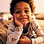 child, smiling, pajamas, pizza_pattern, curly_hair, bed, cozy, indoor, portrait, relaxed, hand_on_face, casual, young, happy, cute, warm_lighting, home, person, sleepwear, resting