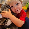 toddler, child, stuffed_animal, hugging, blonde_hair, blue_eyes, indoor, toy, red_shirt, soft_toy, candid, portrait, cute, happy, cozy, furniture, floor, smile, baby, person