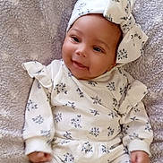 Nayla is registered to the contest to win money with this photo: baby, smiling, infant, headband, bow, white_clothing, floral_pattern, blanket, soft_texture, cute, happy, lying_down, child, young, portrait, cozy, indoors, comfortable, small_hands, face