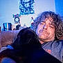 animal, beard, bottle, brown_hair, casual_clothing, closeup, container, couch, cuddling, curly_hair, dog, furniture, home, indoor, living_room, man, pet, portrait, relaxed, wall_art