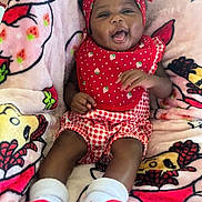 Dasani is registered to the contest to win money with this photo: baby, smiling, infant, headband, red_clothing, bib, blanket, pattern, strawberry, happy, cute, lying_down, socks, child, young, portrait, cozy, soft, colorful, playful