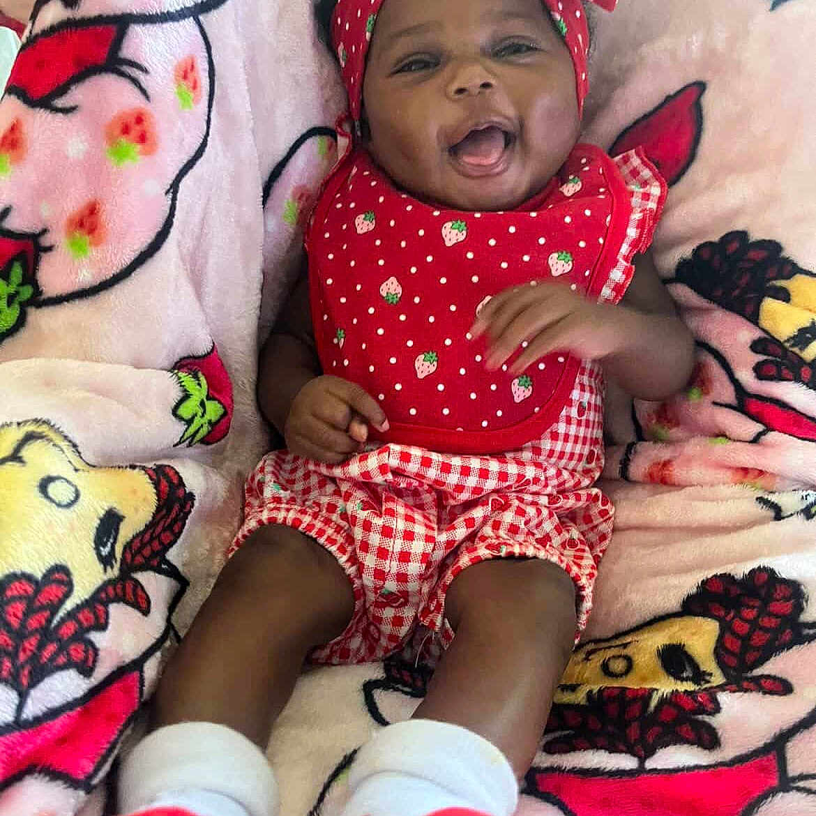 Dasani is registered to the contest to win money with this photo: baby, smiling, infant, headband, red_clothing, bib, blanket, pattern, strawberry, happy, cute, lying_down, socks, child, young, portrait, cozy, soft, colorful, playful