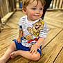 child, toddler, boy, blue_eyes, short_hair, sitting, wooden_deck, casual_clothing, paw_patrol, shirt, shorts, barefoot, outdoor, daylight, smile, curious, cute, portrait, playful, young