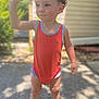 barefoot, blurred_background, casual_clothing, child, curious_expression, cute, daytime, garden, greenery, house_siding, orange_onesie, outdoor, portrait, standing, stone_path, summer, sunlight, toddler, waving_hand, young_child