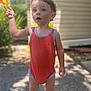 blurry_background, casual_clothing, child, concrete, curious, daylight, expression, face, greenery, onesie, outdoor, person, pointing, short_hair, skin, standing, summer, sunlight, toddler, young_child