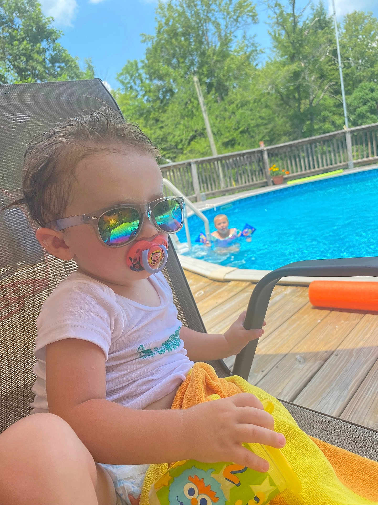 Anthony is registered to the contest to win money with this photo: toddler, child, sunglasses, pacifier, pool, water, swimming, summer, outdoor, sunny, deck, chair, towel, drink_container, reflection, trees, sky, play, relaxation, daytime