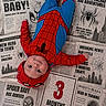 baby, infant, spiderman_costume, costume, newspaper, 3_months, superhero, child, portrait, cute, lying_down, celebration, face, headwear, red, blue, cartoon, spider, web, milestone