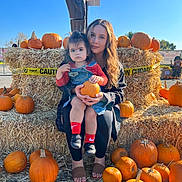 Juliet is registered to the contest to win money with this photo: child, woman, pumpkin, hay_bale, caution_tape, outdoor, fall, autumn, blue_sky, person, sitting, holding, clothing, footwear, nature, daylight, event, smile, cute, family