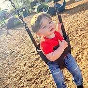 Vinny is registered to the contest to win money with this photo: black_shoes, blue_jeans, casual_clothing, chain, child, daylight, fun, leisure, outdoor, park, play_equipment, playground, recreation, red_shirt, smiling, sunlight, swing, toddler, wood_chips, young_child