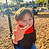 child, toddler, swing, playground, outdoor, sunlight, trees, happy, smiling, red_shirt, blue_jeans, wood_chips, play_equipment, daylight, park, person, casual_clothing, fun, summer, nature