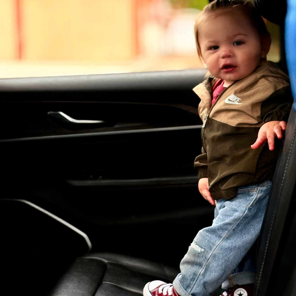 Vinny is registered to the contest to win money with this photo: toddler, child, car_interior, standing, nike_jacket, jeans, sneakers, converse, maroon_shoes, black_leather_seat, window, blurred_background, greenery, yellow_structure, curious_expression, casual_clothing, small_ponytail, indoor, portrait, person