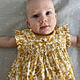 Alma participe au concours pour gagner de l'argent avec cette photo : baby, child, infant, dress, yellow, floral_pattern, smile, face, eyes, arms, blanket, soft_texture, cute, portrait, lying_down, young_child, baby_clothes, human, indoors, closeup