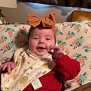 Gwen joined the competition — help win amazing prizes! baby, headband, bow, red_sweater, bib, floral_pattern, couch, indoor, smiling, hand, cute, child, cozy, patterned_fabric, infant, happy, person, seated, warm_lighting, home