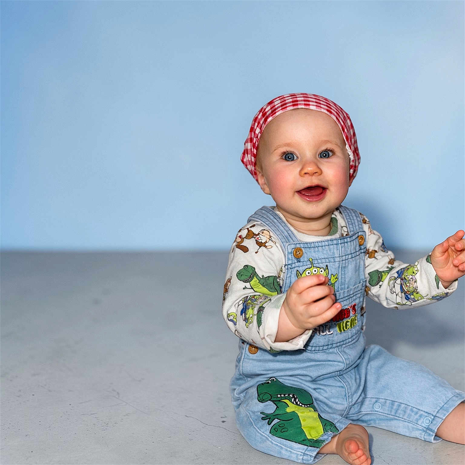 Ambre a rejoint le concours — aidez-le/la à gagner de superbes lots ! baby, infant, toddler, child, smiling, blue_background, denim_overalls, bandana, red_bandana, patterned_shirt, dinosaur_print, sitting, barefoot, studio_floor, portrait, happy, reaching, bright_eyes, headscarf, cute