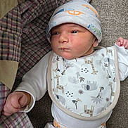 Zane is registered to the contest to win money with this photo: baby, newborn, infant, hat, bib, clothing, plaid_blanket, pattern, animal_print, car_print, face, hand, lying_down, fabric, texture, cute, small, indoors, person, portrait