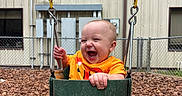 Caspian joined the competition — help win amazing prizes! baby, child, swing, playground, wood_chips, fence, orange_clothing, smiling, happy, outdoor, daylight, seat, metal_chain, building, window, person, joy, fun, infant, park