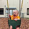 baby, child, swing, playground, wood_chips, fence, orange_clothing, smiling, happy, outdoor, daylight, seat, metal_chain, building, window, person, joy, fun, infant, park