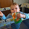 baby, child, toddler, portrait, smiling, happy, eating, food_pouch, snack, baby_food, playpen, nursery, indoor, blue_clothing, reaching, hands, brown_hair, face, stars, wooden_door