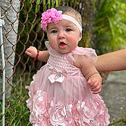 Elianys is registered to the contest to win money with this photo: baby, child, pink_dress, flower_headband, chain_link_fence, outdoor, grass, hand, person, cute, toddler, smiling, portrait, skin, headband, nature, support, young_child, dress, flower