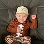 baby, child, smiling, hat, brown_clothing, striped_pants, socks, sitting, couch, indoor, face, person, cute, happy, football_theme, raised_hand, young_child, cozy, casual, portrait