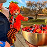 toddler, child, pumpkin_costume, candy, bowl, adult, denim_jacket, pacifier, outdoor, sidewalk, tree, car, costume, autumn, orange, striped_pants, hand, sky, grass, holiday