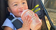 Ryver joined the competition — help win amazing prizes! baby, child, stroller, bottle, drinking, smiling, portrait, outdoor, harness, safety_strap, pink_bow, curly_hair, blue_shirt, infant, face, hands, sunlight, vehicle, seat, happy