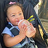 baby, child, stroller, bottle, drinking, smiling, portrait, outdoor, harness, safety_strap, pink_bow, curly_hair, blue_shirt, infant, face, hands, sunlight, vehicle, seat, happy