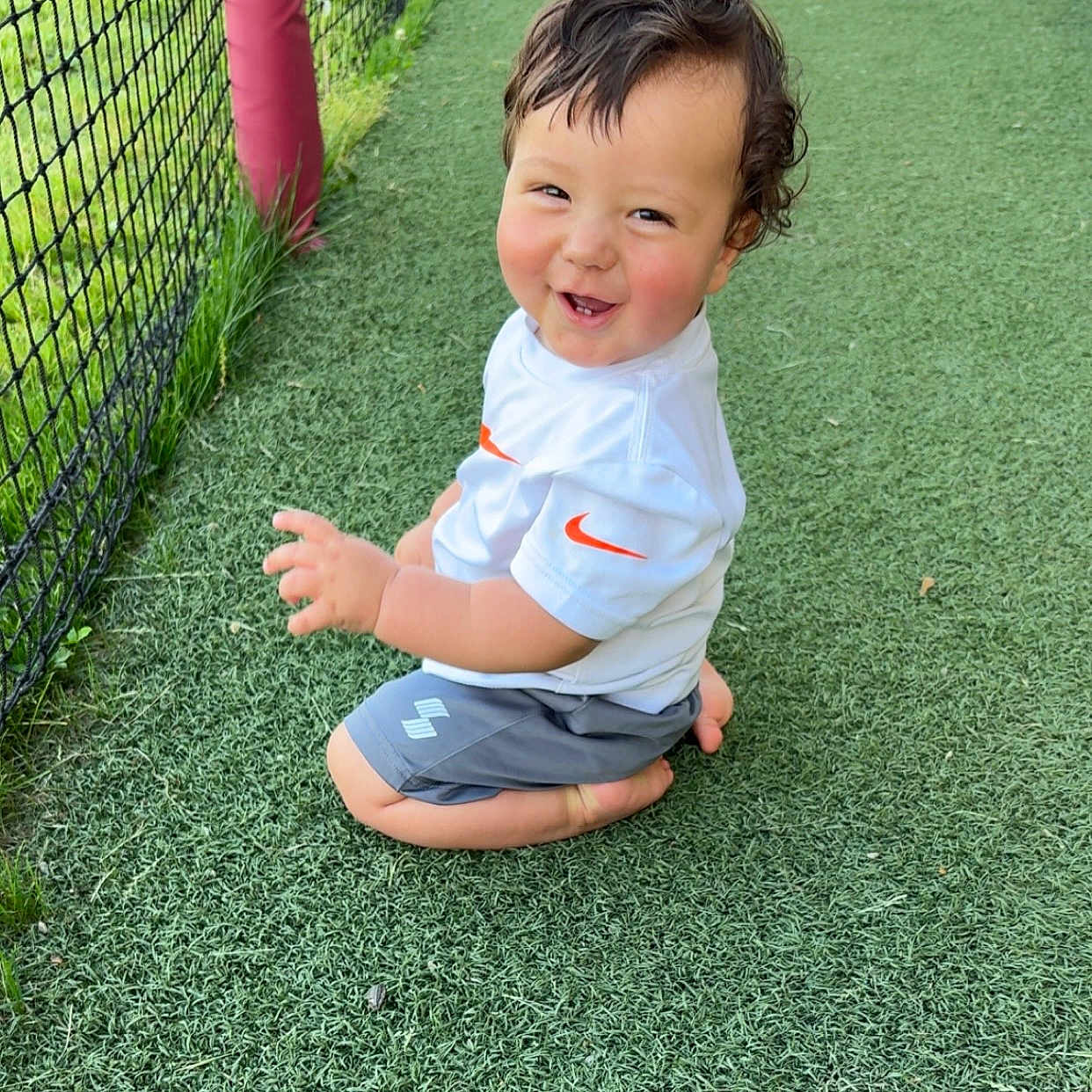 Colden joined the competition — help win amazing prizes! child, toddler, smiling, curly_hair, white_shirt, shorts, kneeling, green_turf, outdoor, net_fence, red_poles, grass, happy, cute, person, playing, young, sunny, daylight, joy