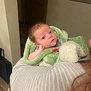 Ron joined the competition — help win amazing prizes! baby, infant, blanket, green_blanket, person, indoor, holding, face, eyes, curious, soft_texture, warm_lighting, wood_floor, striped_clothing, hand, head, portrait, newborn, cozy, human