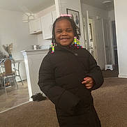 Imani joined the competition — help win amazing prizes! carpet, child, clothing, colorful_beads, cozy, door, furniture, hair, happy, home, indoor, kitchen, light, person, portrait, puffer_jacket, room, smiling, standing, wall