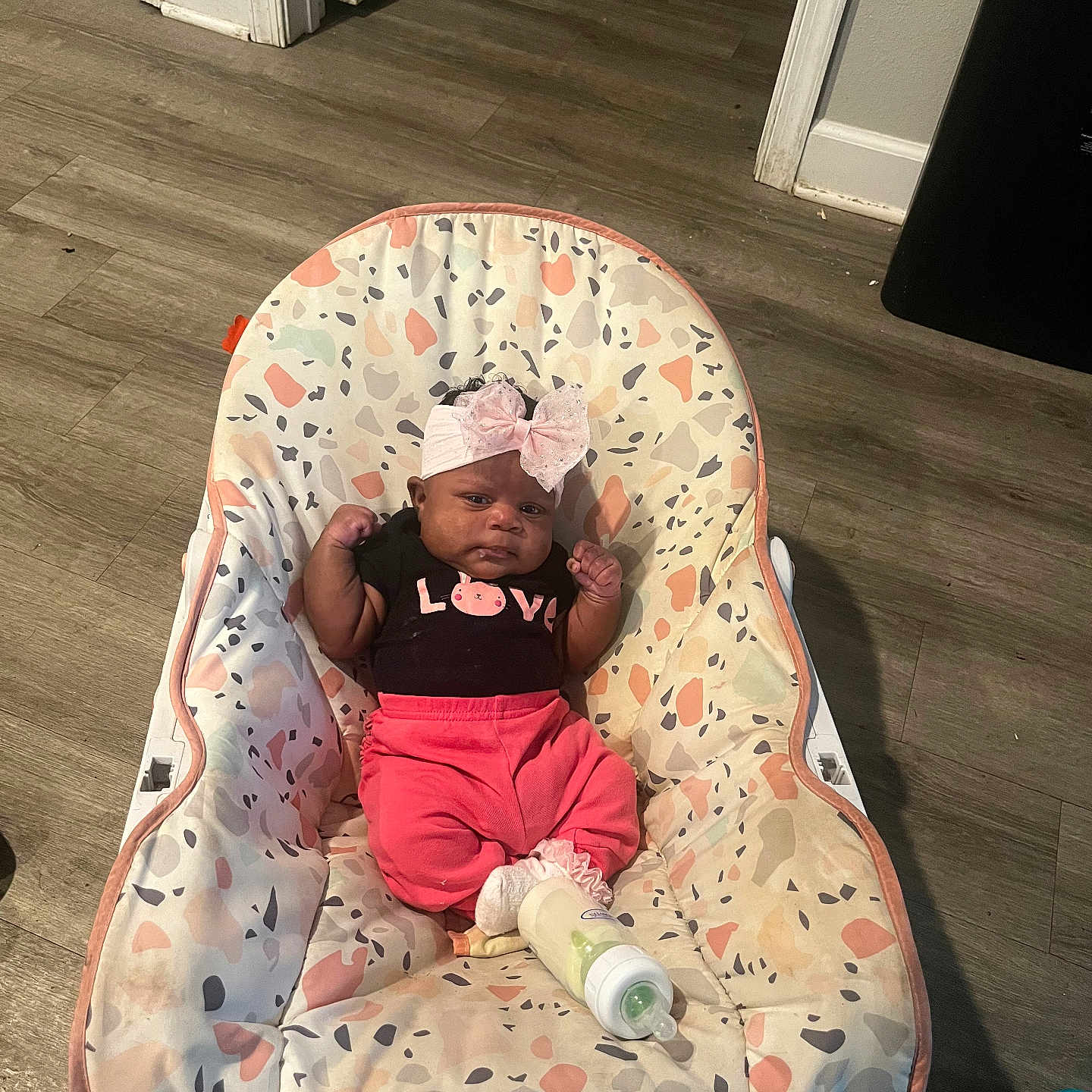 Jhream is registered to the contest to win money with this photo: baby, infant, child, portrait, sitting, bouncer, baby_chair, pink_bow, headband, clothing, black_shirt, pink_pants, bottle, indoor, home, wood_floor, doorway, furniture, awake, serious_expression