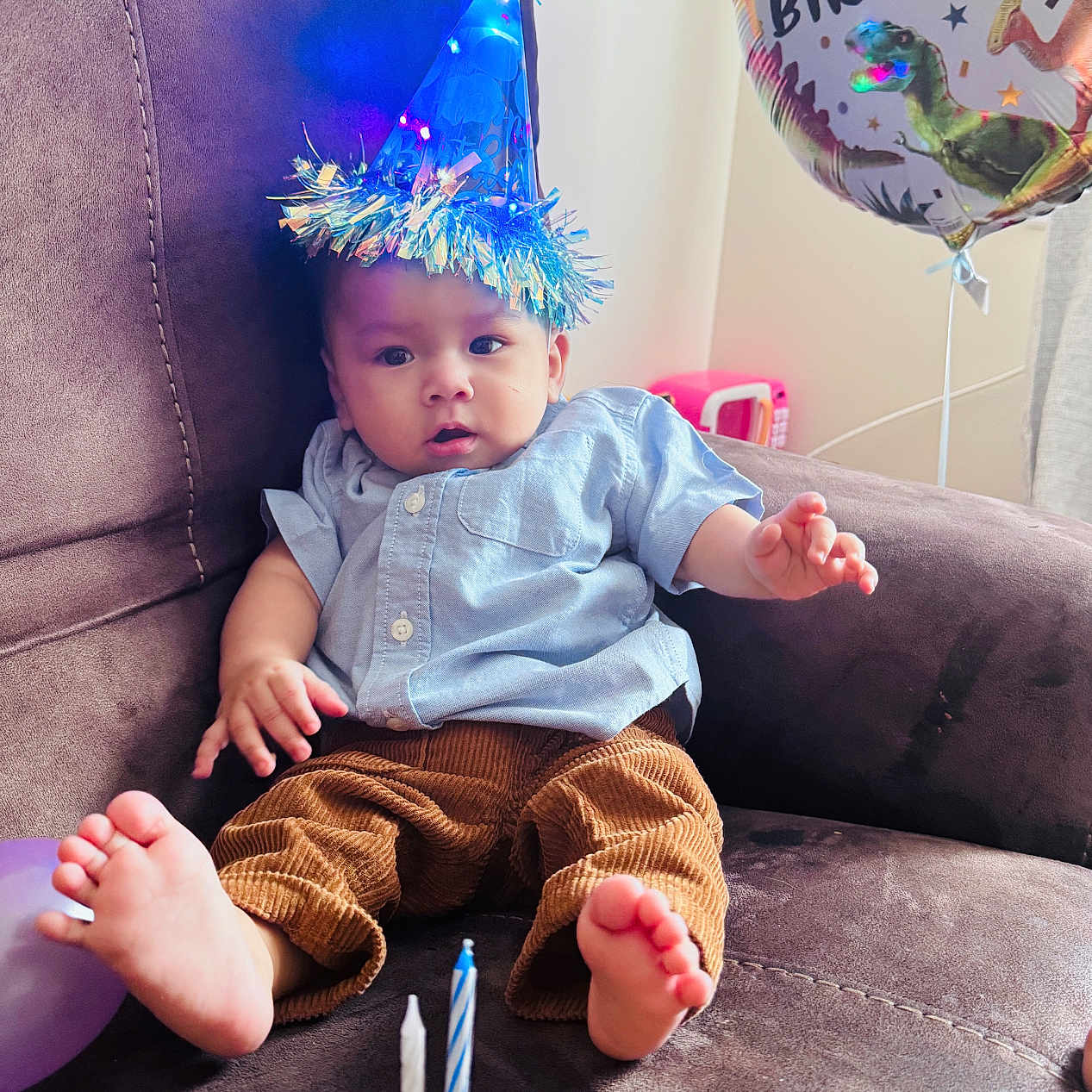 Jilson is registered to the contest to win money with this photo: baby, balloon, birthday, blue_shirt, brown_pants, candles, celebration, child, chocolate, clothing, couch, curious, dinosaur, donut, feet, festive, home, indoors, party_hat, relaxed