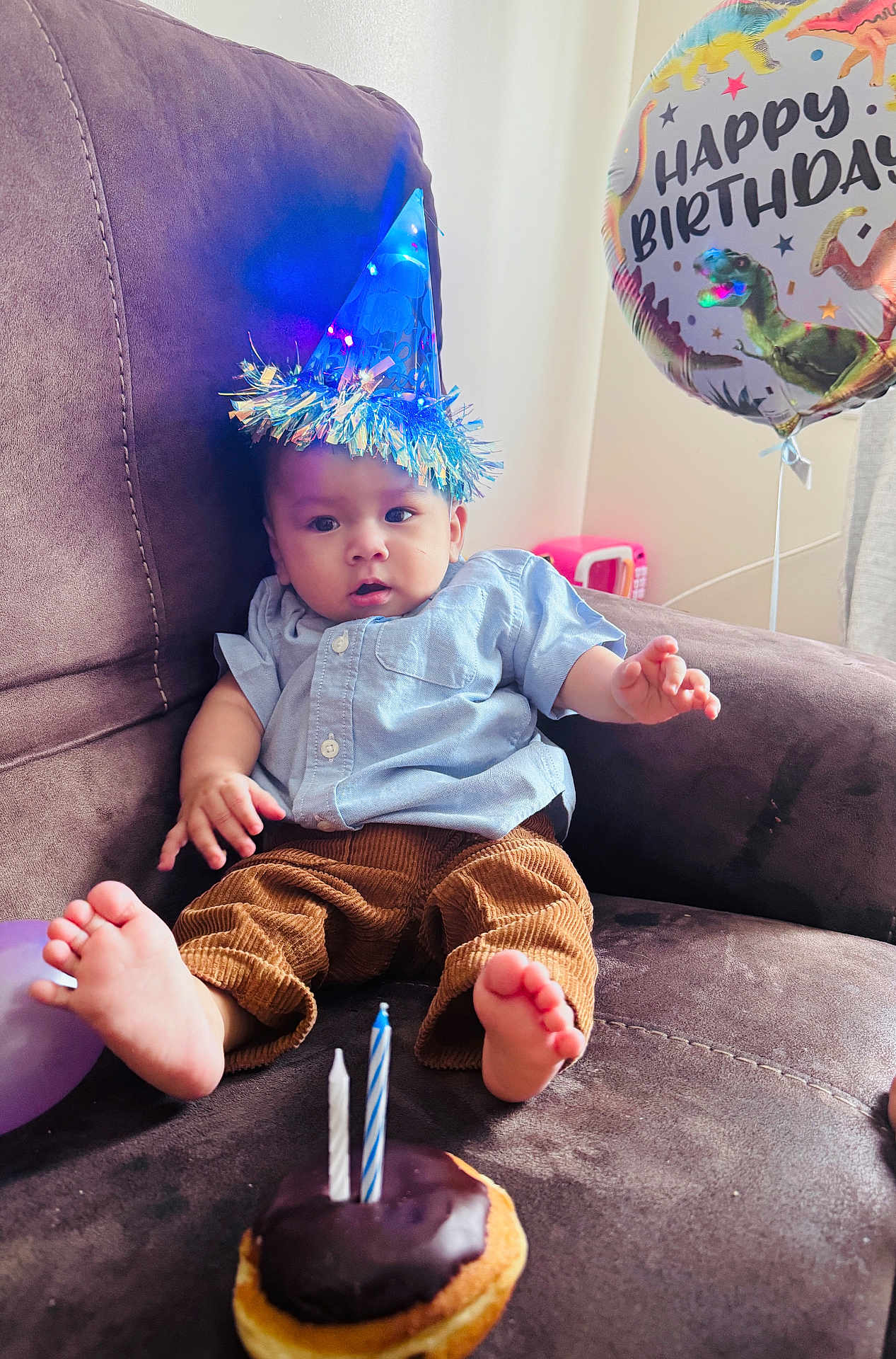 Jilson is registered to the contest to win money with this photo: baby, party_hat, birthday, couch, donut, chocolate, candles, balloon, dinosaur, celebration, indoors, child, curious, clothing, blue_shirt, brown_pants, feet, relaxed, festive, home