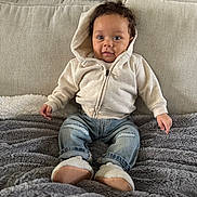 Lavonte is registered to the contest to win money with this photo: baby, hoodie, jeans, blanket, couch, indoor, child, smiling, sitting, curly_hair, footwear, plush, cozy, relaxed, portrait, furniture, living_room, casual_clothing, young_child, television