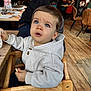 toddler, child, high_chair, hoodie, blue_eyes, bread, cup, wooden_chair, indoor, restaurant, people, table, menu, flooring, casual_clothing, curious, seated, daylight, person, furniture