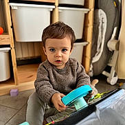 Gabriel a rejoint le concours — aidez-le/la à gagner de superbes lots ! child, toddler, toy, steering_wheel, sweater, blue_eyes, indoor, floor, storage_bins, baby_gear, baby_rocker, playing, curious, person, furniture, cozy, young_child, brown_hair, expression, home