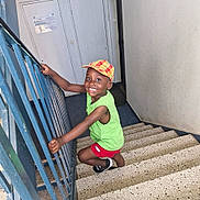 Waly participe au concours pour gagner de l'argent avec cette photo : child, smiling, stairs, cap, green_shirt, red_shorts, indoor, railing, happy, person, footwear, climbing, young_child, portrait, casual_clothing, boy, playful, bright_colors, concrete_stairs, handrail