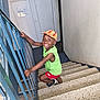child, smiling, stairs, cap, green_shirt, red_shorts, indoor, railing, happy, person, footwear, climbing, young_child, portrait, casual_clothing, boy, playful, bright_colors, concrete_stairs, handrail
