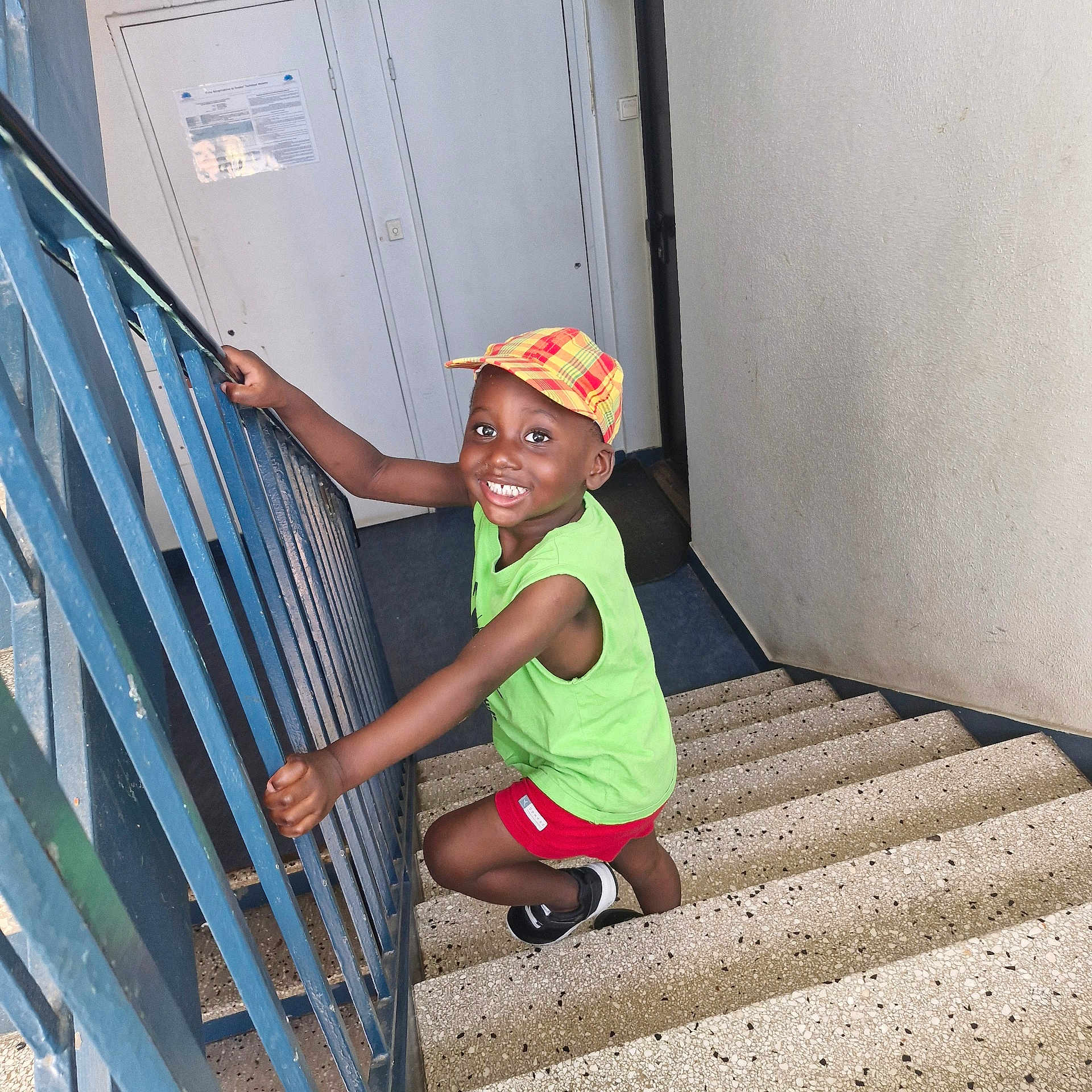 Waly participe au concours pour gagner de l'argent avec cette photo : child, smiling, stairs, cap, green_shirt, red_shorts, indoor, railing, happy, person, footwear, climbing, young_child, portrait, casual_clothing, boy, playful, bright_colors, concrete_stairs, handrail