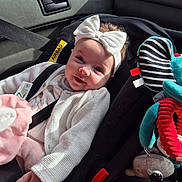 Isabella is registered to the contest to win money with this photo: baby, child, car_seat, headband, bow, smile, plush_toy, seatbelt, car_interior, sunlight, white_clothing, infant, toy, happy, cute, safety, vehicle, seat, child_safety, portrait