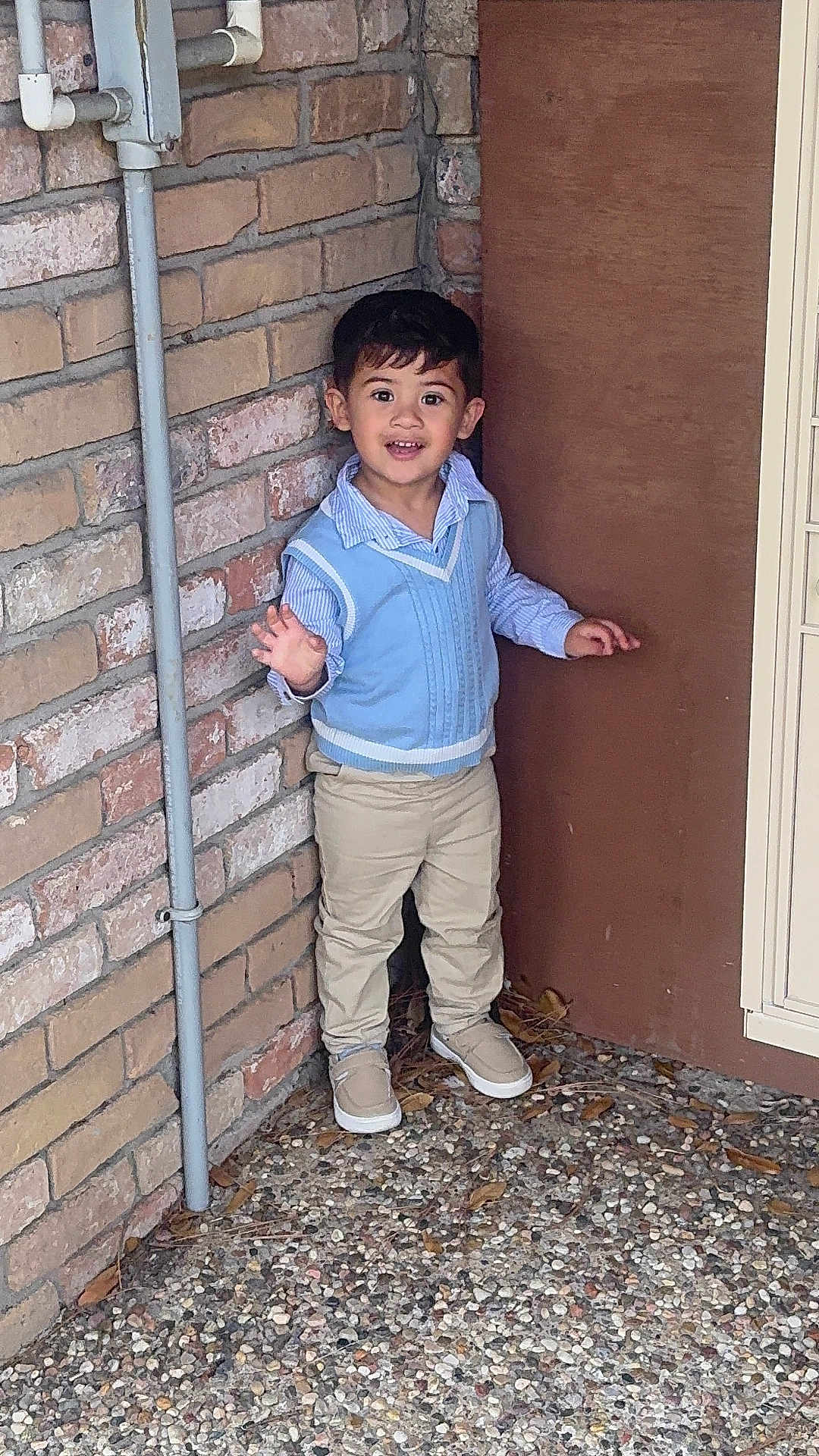 Liam joined the competition — help win amazing prizes! child, boy, smiling, waving, blue_sweater_vest, striped_shirt, beige_pants, shoes, brick_wall, wooden_panel, corner, outdoor, standing, happy, cute, person, casual, daylight, young, portrait