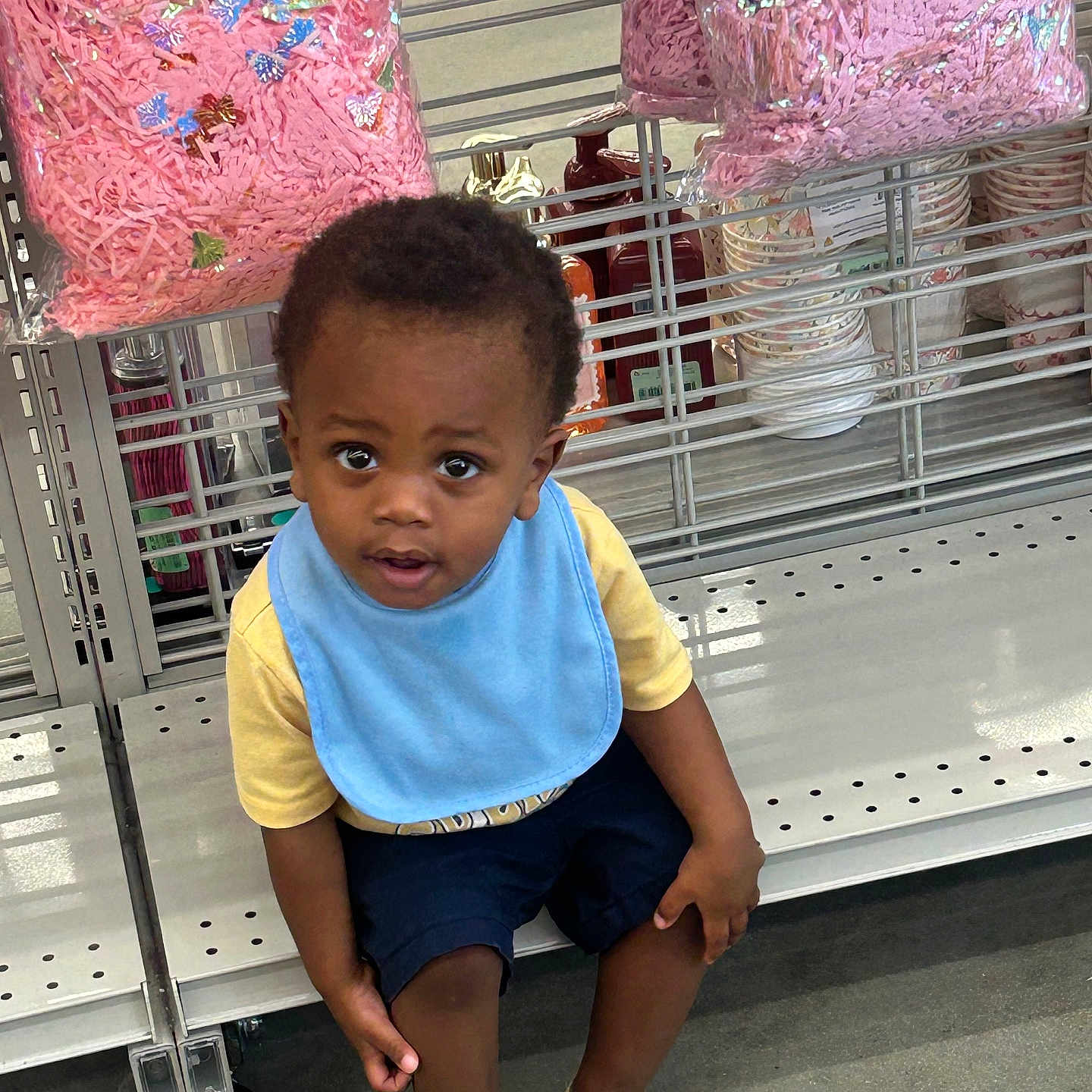 Jaceyon is registered to the contest to win money with this photo: child, toddler, boy, sitting, face, portrait, curious, indoors, store, shopping, retail, shelf, craft_supplies, pink, gift_bags, bib, yellow_shirt, blue_clothing, sandals, floor