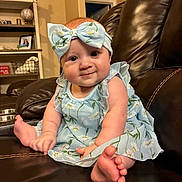 Blakeley is registered to the contest to win money with this photo: baby, child, infant, dress, headband, floral_pattern, blue, smile, leather_couch, living_room, furniture, person, foot, hand, indoors, sitting, cute, portrait, happy, home