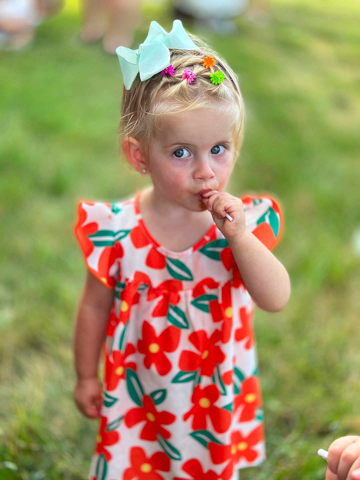 Daniella is registered to the contest to win money with this photo: child, girl, lollipop, hair_bow, hair_clips, blue_eyes, floral_dress, outdoor, grass, summer, cute, portrait, happy, colorful, young_child, fashion, playful, person, sweet, fun